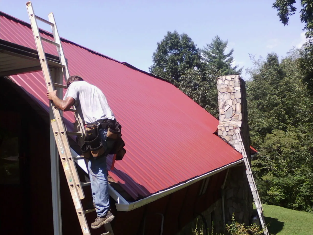 Roofer on a ladder beside a red metal roof and stone chimney — Soffit & Fascia Repair by Roofing Brothers in Double Springs